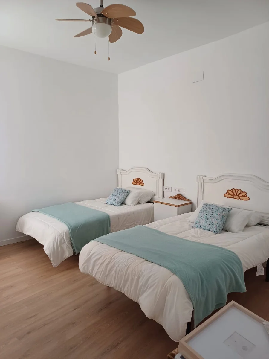 Dormitorio recién reformado con dos camas hechas, ventilador de techo y suelo de tarima.