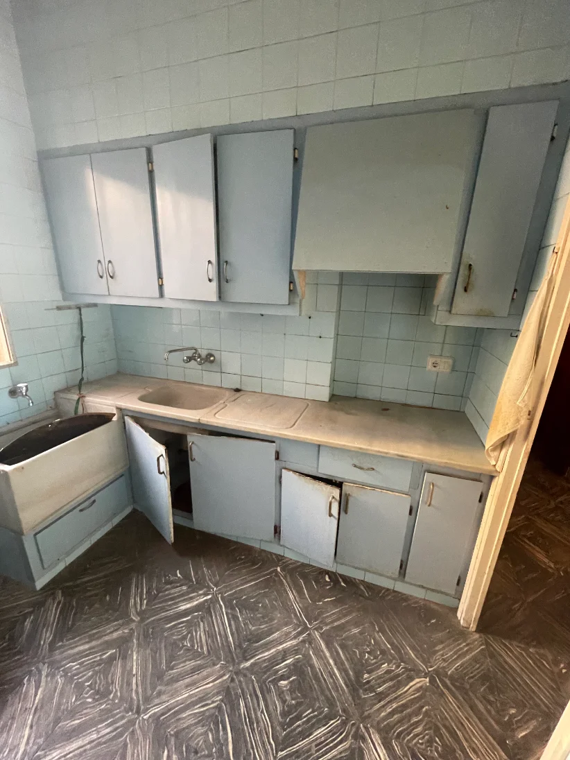 Cocina antes de la reforma: muebles de madera pintados de azul de los años setenta, alicatado antiguo y suelo deteriorado.
